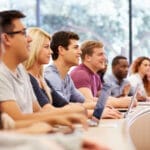 A row of students in lecture call listening to professor with laptops